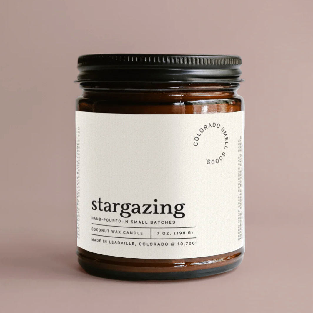 A 7 oz coconut wax candle in an amber glass jar with a black metal lid and a white paper label. The label features the scent name 'stargazing' and the brand 'Colorado Small Batch' in black text, set against a solid taupe background.