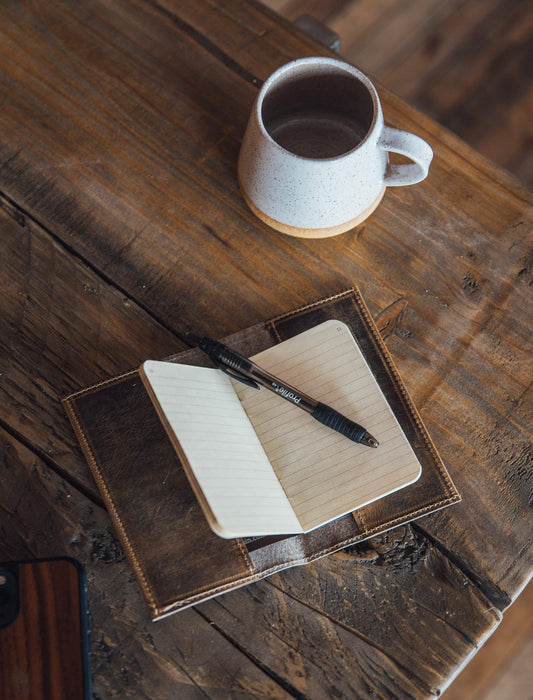 Leather notebook cover with beige notebook, black pen, and white handmade coffee mug