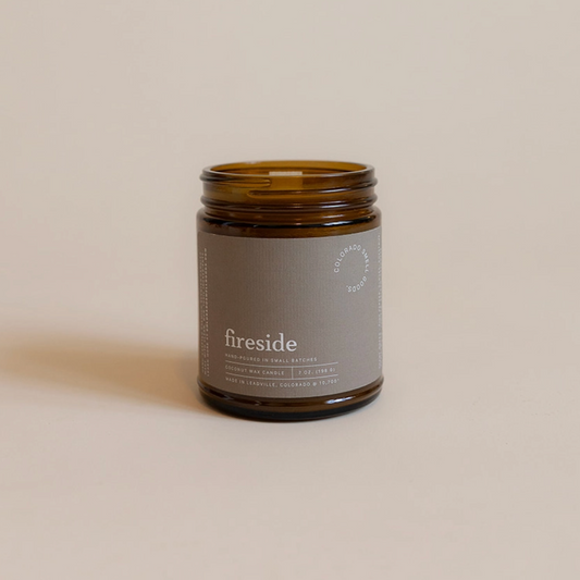 An amber glass jar candle with a gray label reading 'fireside' and 'FIREPLACE CANDLES' on a warm beige background.