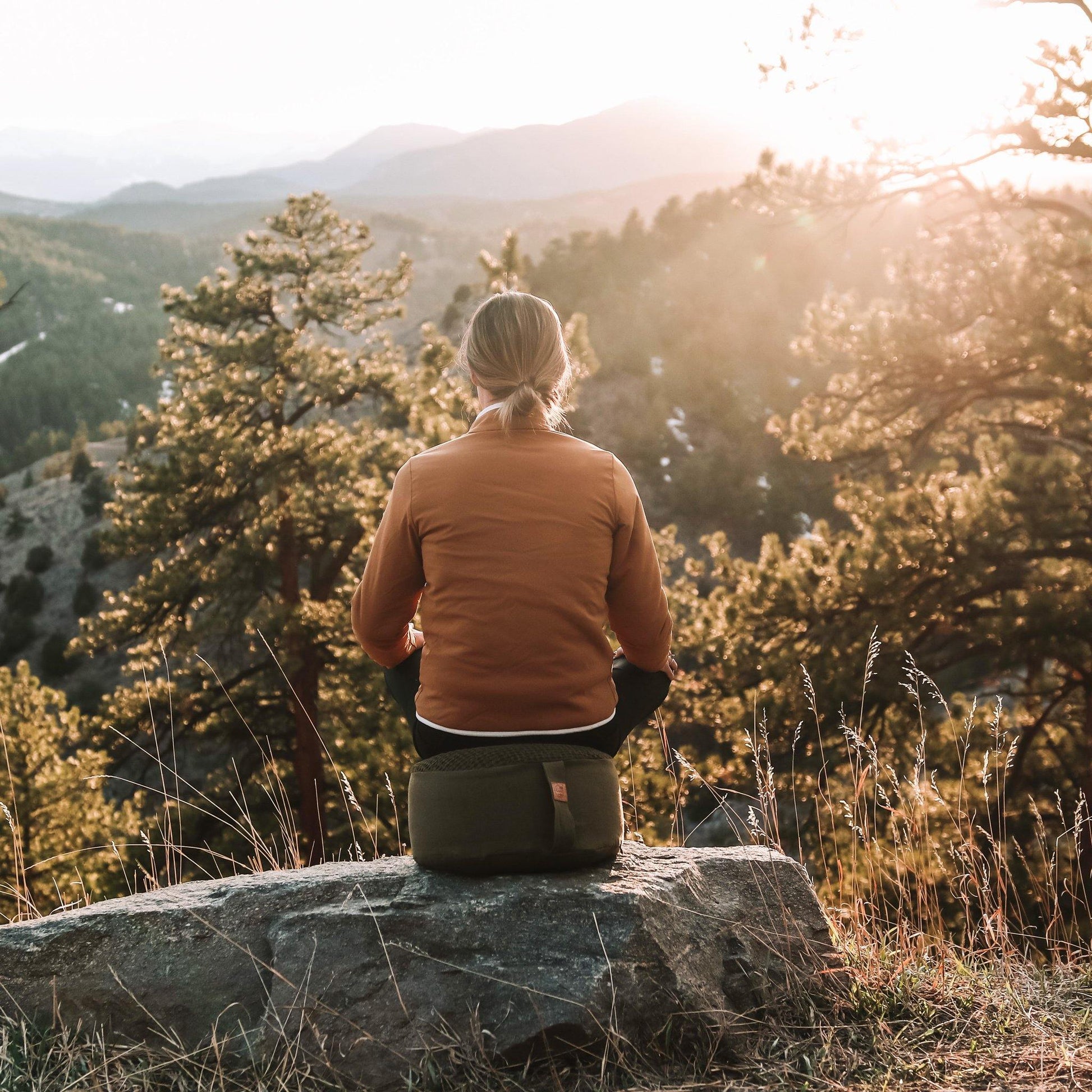 Woman meditating on an olive-colored zafu cushion with a beautiful mountain sunset in Colorado